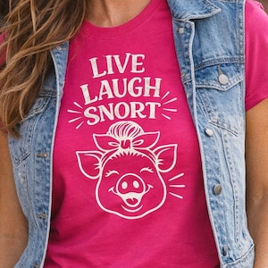 May include: A hot pink t-shirt with the words "LIVE LAUGH SNORT" and a pig graphic. The shirt is worn with a denim vest, blue jeans, a brown belt, and a straw hat. The person is smiling.