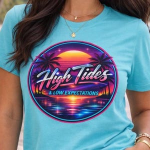 May include: A light blue t-shirt with a round graphic featuring palm trees, a sunset, and the words "High Tides & Low Expectations" in a retro font. The shirt has short sleeves and a crew neck.