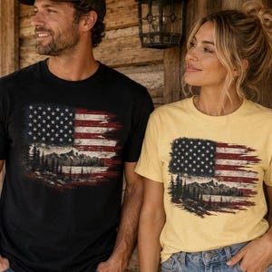 Couple wearing black and butter yellow patriotic mountain flag graphic Comfort Colors t shirts outside rustic wooden cabin with western casual styling