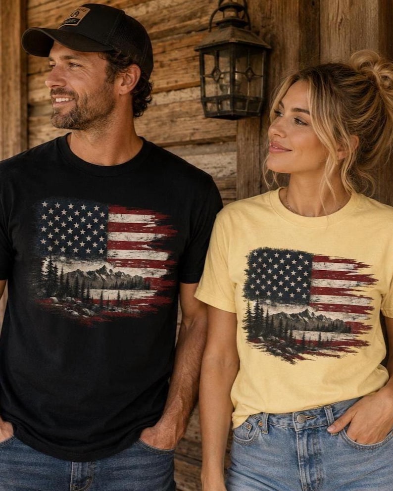 Couple wearing black and butter yellow patriotic mountain flag graphic Comfort Colors t shirts outside rustic wooden cabin with western casual styling