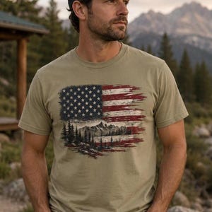 Man wearing patriotic wilderness flag graphic Comfort Colors t shirt near mountain river cabin with baseball hat and jeans