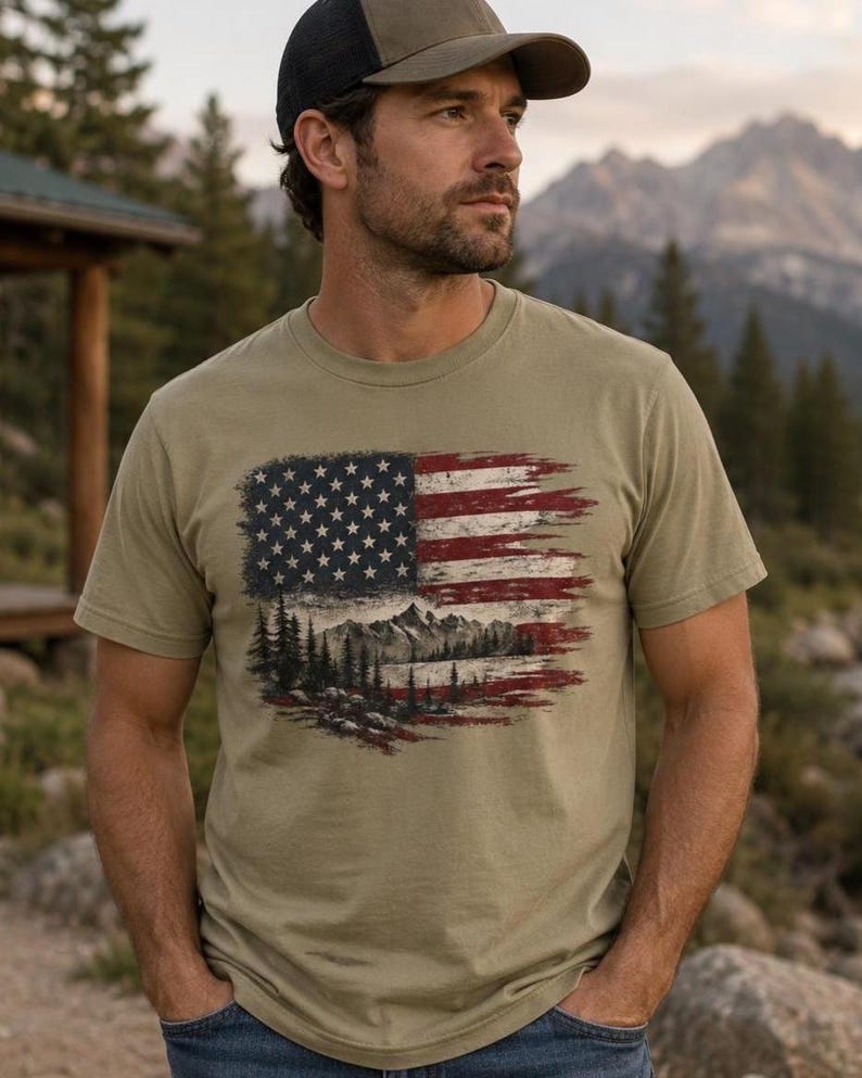 Man wearing patriotic wilderness flag graphic Comfort Colors t shirt near mountain river cabin with baseball hat and jeans