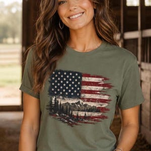Woman wearing moss patriotic mountain flag graphic Comfort Colors t shirt inside rustic barn with light wash jeans western bracelets and front tuck styling