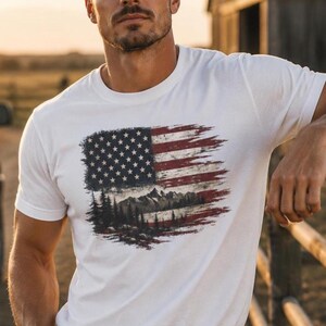 Handsome man wearing white patriotic wilderness flag graphic Comfort Colors t shirt on ranch fence at sunset with jeans and western belt