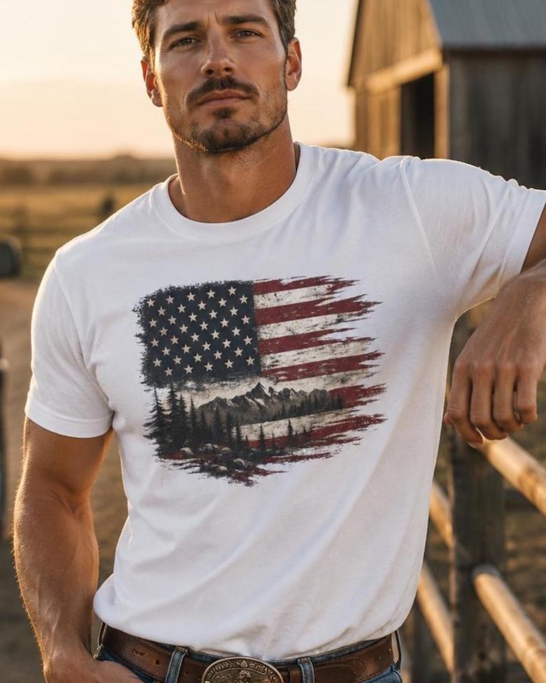 Handsome man wearing white patriotic wilderness flag graphic Comfort Colors t shirt on ranch fence at sunset with jeans and western belt