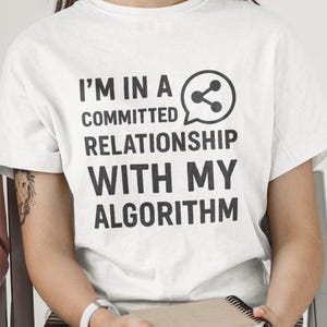 May include: White t-shirt with the text "I'M IN A COMMITTED RELATIONSHIP WITH MY ALGORITHM" in black. The shirt features a speech bubble with a share icon. The person is holding a stack of notebooks.