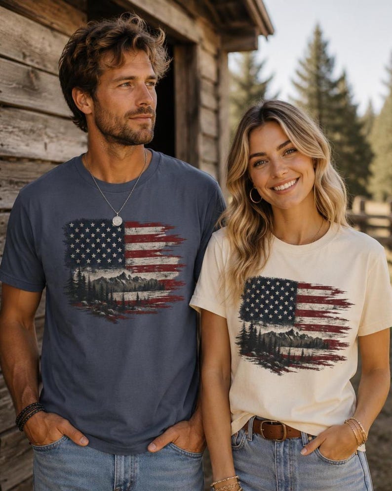 Couple wearing navy blue and soft cream patriotic mountain flag graphic Comfort Colors t shirts beside rustic cabin in outdoor western setting