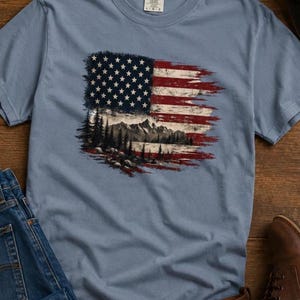 Patriotic mountain flag graphic Comfort Colors t shirt flat lay with jeans leather boots black hat and rustic wood background