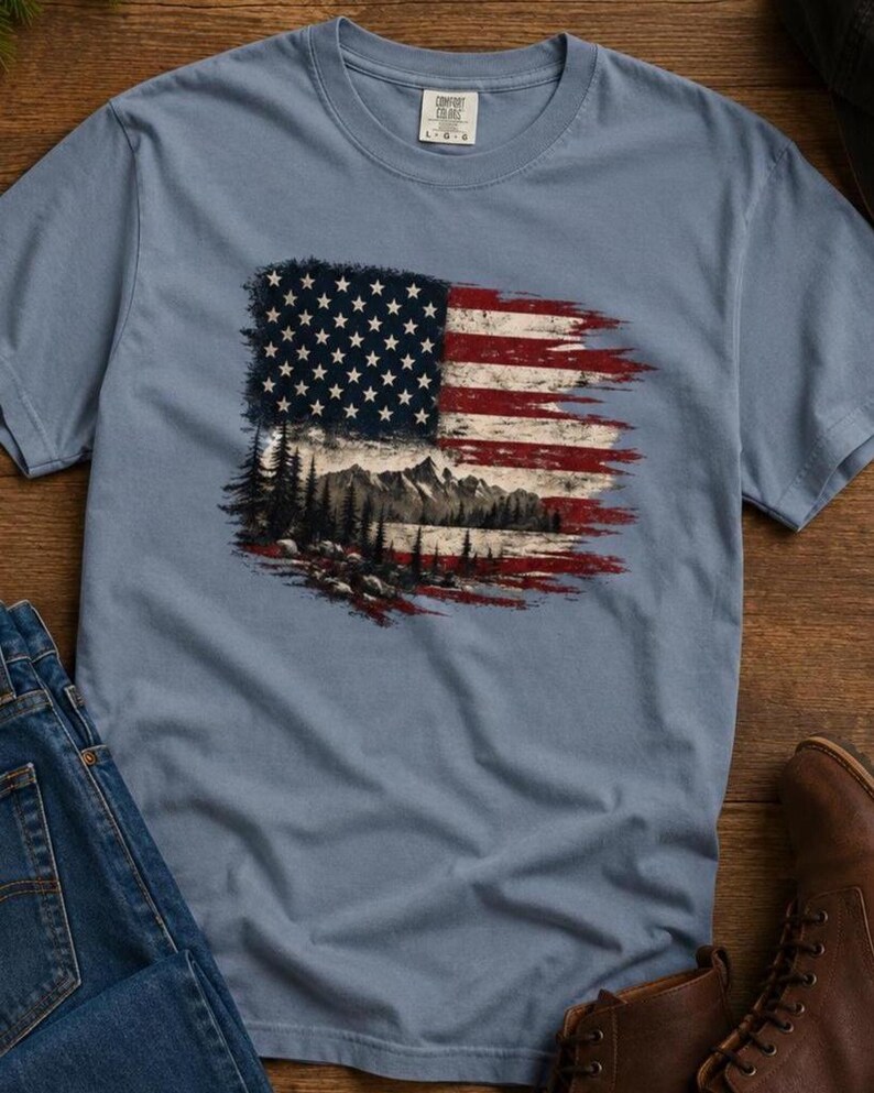 Patriotic mountain flag graphic Comfort Colors t shirt flat lay with jeans leather boots black hat and rustic wood background