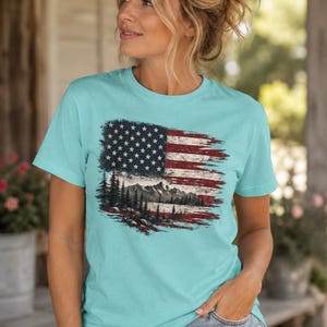 Woman wearing patriotic mountain flag graphic Comfort Colors t shirt with distressed denim shorts on farmhouse porch