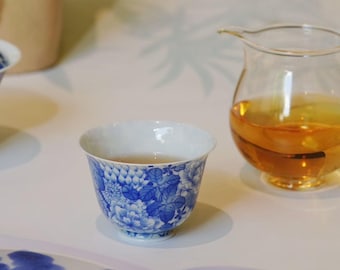 May include: A white and blue floral teacup filled with tea, next to a glass pitcher of tea. The teacup has a blue floral pattern on the outside and a white interior. The tea is amber-colored.