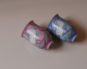 May include: Two ceramic vases, one lavender and one blue, each adorned with a floral design. The vases feature a flared rim and a rounded body, with a delicate pattern of white flowers and green leaves.