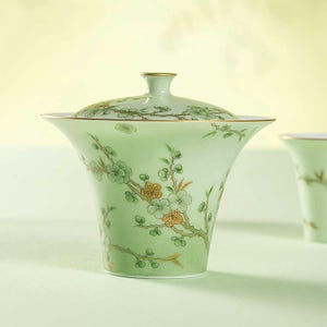 May include: A pale green porcelain tea cup with a matching lid. The cup and lid are decorated with a floral design in shades of green and gold. The cup has a flared shape and a gold rim.