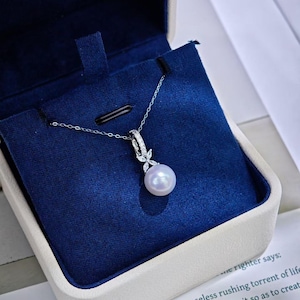 May include: A delicate silver necklace with a large, round pearl pendant, displayed in a blue velvet jewelry box. The pendant is accented with small, sparkling stones. The box is white with a blue interior.