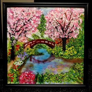Peut inclure: Une œuvre d'art encadrée représentant une scène de jardin sereine. Le tableau montre un pont rouge au-dessus d'un ruisseau bleu, entouré d'un feuillage vert luxuriant et de cerisiers en fleurs roses vibrants. L'œuvre est dans un cadre noir.