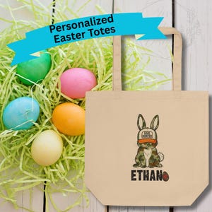 May include: A beige tote bag with a cartoon bunny wearing a hat that says "Egg Hunter" and the name "Ethan" below. The bag is next to colorful Easter eggs in green paper grass. Text on a blue banner reads "Personalized Easter Totes."
