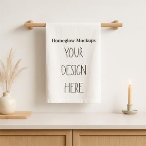 Może przedstawiać: Biały ręcznik z napisem "Homeglow Mockups YOUR DESIGN HERE" zawieszony na drewnianym drążku. Zapalona świeca w beżowym świeczniku i wazon z suszonymi roślinami znajdują się na jasnej powierzchni.