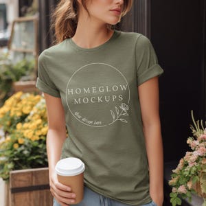 May include: Olive green t-shirt with rolled sleeves. The shirt features a white circular logo with the text "HOMEGLOW MOCKUPS" and "Your design here". A person is holding a brown paper coffee cup with a white lid.