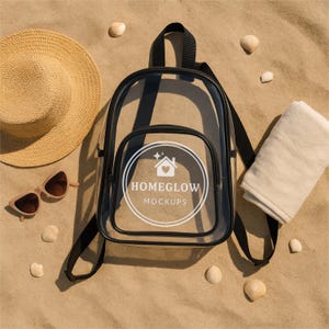 May include: A clear backpack with black trim, a straw hat, sunglasses, and a folded towel are arranged on a sandy surface. The backpack has a white logo that says "HOMEGLOW MOCKUPS". Several seashells are scattered around the items.