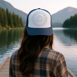 Könnte beinhalten: Eine marineblaue und weiße Trucker-Mütze mit dem Text "YOUR DESIGN HERE" und "HOMEGLOW MOCKUPS", der eine Hutgrafik umgibt. Die Mütze wird von einer Person mit langen braunen Haaren getragen, vor dem Hintergrund eines Sees und Bergen.