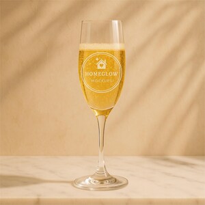 Può includere: Un calice da champagne pieno di liquido dorato frizzante. Il bicchiere ha un logo che recita "HOMEGLOW MOCKUPS" in un design circolare. Il bicchiere poggia su una superficie di marmo bianco su uno sfondo neutro.