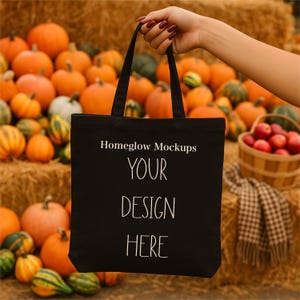 Könnte beinhalten: Eine schwarze Tragetasche mit dem Text "Homeglow Mockups YOUR DESIGN HERE" wird vor einem Hintergrund aus Kürbissen und Heuballen gehalten. Die Tasche hat zwei schwarze Griffe und ist aus robustem Material gefertigt.