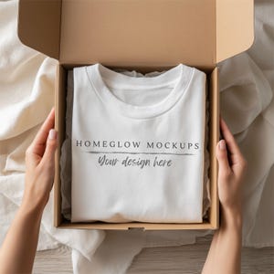 May include: A white t-shirt with the text "HOMEGLOW MOCKUPS" and "Your design here" is presented inside a brown cardboard box. The box is open, revealing the shirt neatly folded. The scene is set on a light-colored fabric.