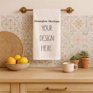 May include: A kitchen scene with a white towel hanging on a brass rod, featuring the text "Homeglow Mockups YOUR DESIGN HERE." A woven placemat, a bowl of oranges and lemons, a mug, and potted plants are on a wooden countertop.