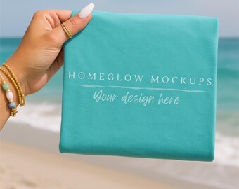 May include: A folded turquoise fabric with the text "HOMEGLOW MOCKUPS" and "Your design here" in white script. The fabric is held by a hand with a gold ring and bracelet, with a beach and ocean background.