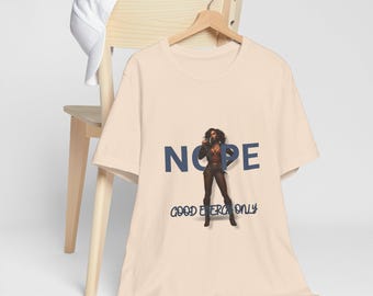 May include: A light beige t-shirt with the word "NOPE" in navy blue block letters and a graphic of a woman. Below the word "NOPE" is the text "GOOD ENERGY ONLY". A white baseball cap rests on a wooden chair.
