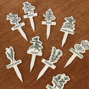 May include: A selection of white wooden herb garden markers featuring black illustrations and names like basil and parsley. The markers are displayed on a brown surface. Each marker is labelled with the herb's name.
