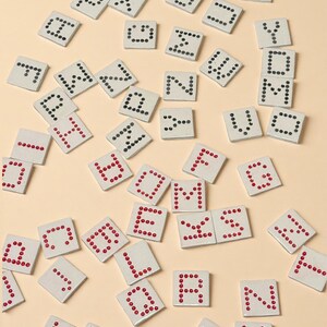 May include: Scattered white square tiles with letters formed by red or black dots. The tiles are arranged on a light beige surface, creating a playful and educational visual. The letters are in various fonts.