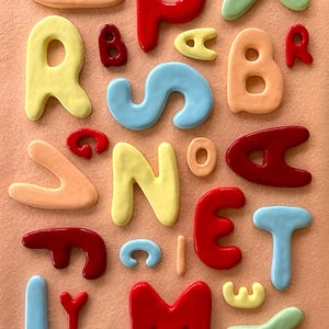 May include: A selection of brightly coloured, glossy, three-dimensional letters in various hues, including red, yellow, blue, and green, arranged on a peach-coloured background. The letters appear to be made of a smooth, possibly ceramic, material.