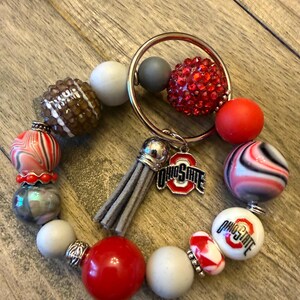 May include: A circular keychain featuring a mix of beads in red, white, gray, and black, including a football-textured bead, a red rhinestone bead, and two beads with the Ohio State logo. A gray suede tassel and a metal Ohio State charm are attached.