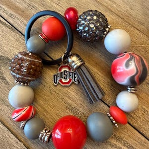May include: A beaded bracelet featuring a variety of beads in red, gray, white, and brown. The bracelet includes a charm with the Ohio State logo and a gray tassel. The beads are of various shapes and sizes, including round, oval, and crystal-studded.