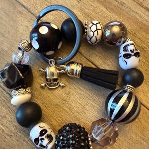 May include: A beaded keychain featuring a dark metal ring, a silver skull charm, and a black suede tassel. The beads include black and white polka dots, matte black spheres, white skull beads, striped spheres, and faceted black crystals.