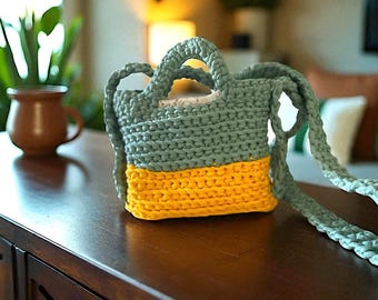 May include: A two-tone crochet bag, with a muted green upper section and a bright yellow base. It features a short handle and a long strap. The bag is displayed on a dark wooden surface. The bag's design suggests it is handmade.