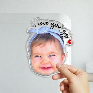 May include: A custom acrylic photo magnet featuring a smiling baby wearing a light blue headband. The text "i love you, gigi" is printed in black script above the baby's head, accompanied by a small red heart icon on the right side.