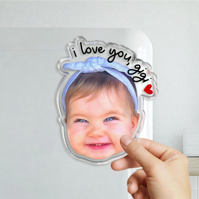 May include: A custom acrylic photo magnet featuring a smiling baby wearing a light blue headband. The text "i love you, gigi" is printed in black script above the baby's head, accompanied by a small red heart icon on the right side.