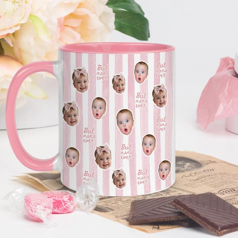 May include: A ceramic mug with a pink and white vertical striped pattern, featuring repeated photographic portraits of a baby wearing a pink bow and the repeating text Best nana ever in a cursive script, with a solid pink handle and interior.