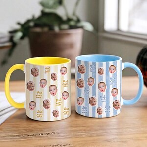 May include: Two ceramic mugs featuring a repeating pattern of a baby's face and the text "Best papa ever" or "Best dad ever". The left mug has a yellow handle and interior with yellow stripes, while the right mug has a light blue handle and interior with blue stripes.