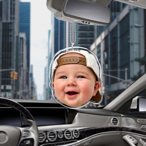 Puede incluir: Un adorno colgante para coche de acr&iacute;lico transparente con un retrato en alta resoluci&oacute;n de un beb&eacute; sonriente que lleva una gorra de b&eacute;isbol blanca con visera marr&oacute;n. El adorno est&aacute; suspendido por una cadena de bolas de metal plateado.