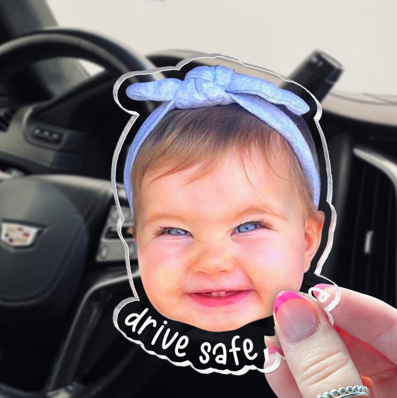 Puede incluir: Un adorno de acr&iacute;lico transparente con una impresi&oacute;n fotogr&aacute;fica de un beb&eacute; sonriente que lleva una diadema azul claro. El texto "drive safe" est&aacute; impreso en blanco en la parte inferior del recorte. El adorno tiene un borde liso y acabado brillante.