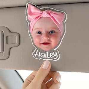 Puede incluir: Un clip para parasol de coche de acr&iacute;lico personalizado con una fotograf&iacute;a de alta resoluci&oacute;n de un beb&eacute; con una diadema de lazo rosa claro. El recorte de acr&iacute;lico transparente tiene la forma de la cabeza del beb&eacute; y el nombre Hailey en blanco.