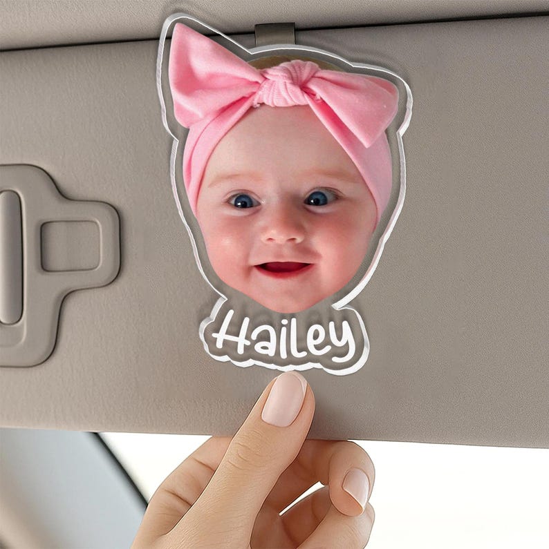 Puede incluir: Un clip para parasol de coche de acr&iacute;lico personalizado con una fotograf&iacute;a de alta resoluci&oacute;n de un beb&eacute; con una diadema de lazo rosa claro. El recorte de acr&iacute;lico transparente tiene la forma de la cabeza del beb&eacute; y el nombre Hailey en blanco.