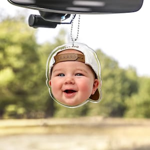 Puede incluir: Un adorno colgante para coche de acr&iacute;lico transparente con una imagen impresa de alta resoluci&oacute;n de un beb&eacute; sonriente que lleva una gorra de b&eacute;isbol blanca con visera marr&oacute;n. El adorno cuelga de una cadena de bolas de metal plateado.