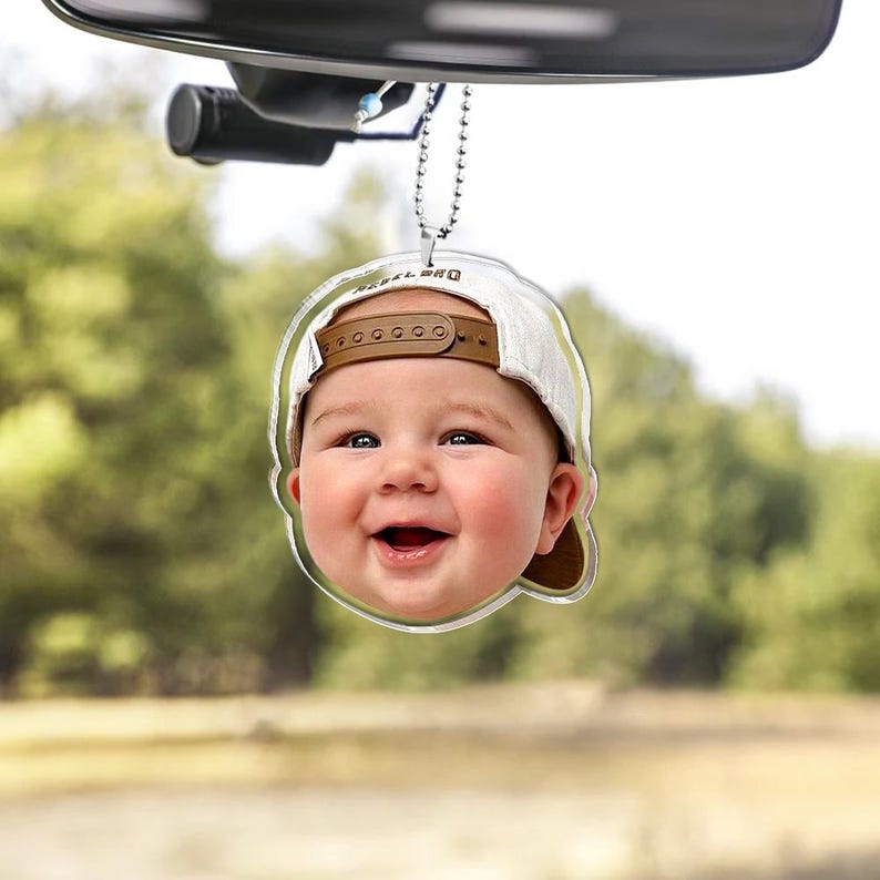 Puede incluir: Un adorno colgante para coche de acr&iacute;lico transparente con una imagen impresa de alta resoluci&oacute;n de un beb&eacute; sonriente que lleva una gorra de b&eacute;isbol blanca con visera marr&oacute;n. El adorno cuelga de una cadena de bolas de metal plateado.
