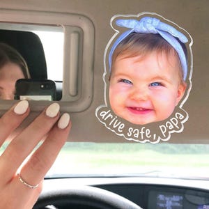 Puede incluir: Un clip de acr&iacute;lico transparente para el parasol del coche con una fotograf&iacute;a de alta resoluci&oacute;n de un beb&eacute; sonriente con una diadema azul claro. La forma recortada incluye el texto blanco "drive safe, papa" en letra cursiva.