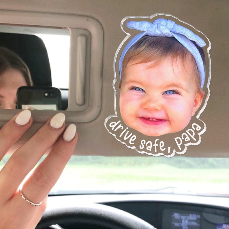 Puede incluir: Un clip de acr&iacute;lico transparente para el parasol del coche con una fotograf&iacute;a de alta resoluci&oacute;n de un beb&eacute; sonriente con una diadema azul claro. La forma recortada incluye el texto blanco "drive safe, papa" en letra cursiva.