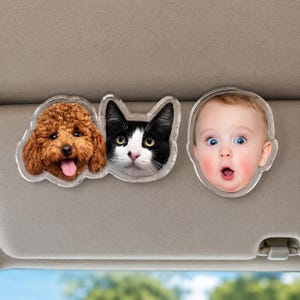 Puede incluir: Dos clips de acr&iacute;lico personalizados para visera de coche con retratos fotogr&aacute;ficos. Un clip muestra un perro marr&oacute;n de pelo rizado y un gato blanco y negro juntos. El segundo clip presenta el retrato de un beb&eacute; con expresi&oacute;n de sorpresa.